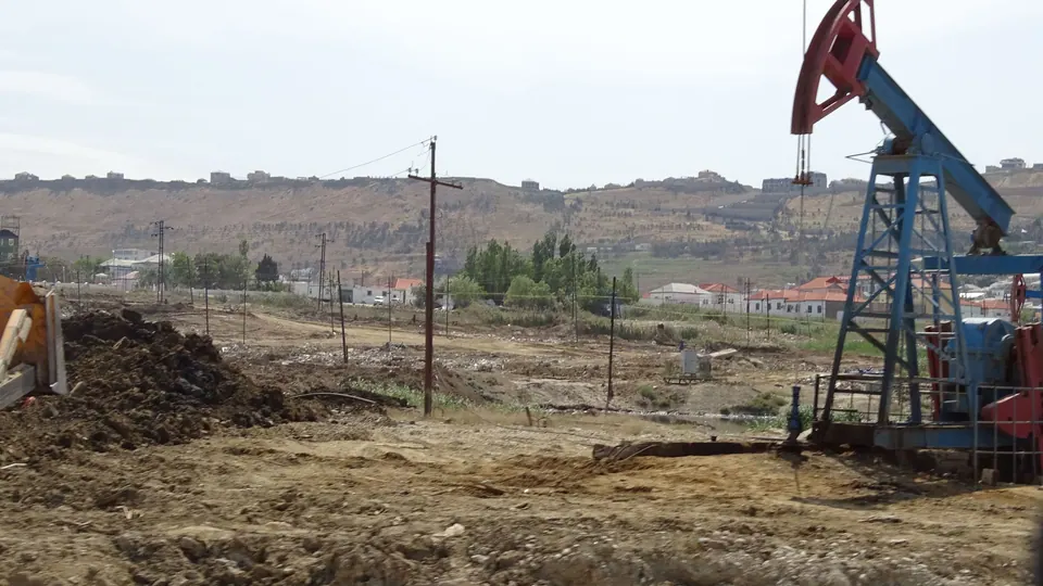 A blue and red pumpjack beside bare ground and utility poles, with houses on a ridge behind.
