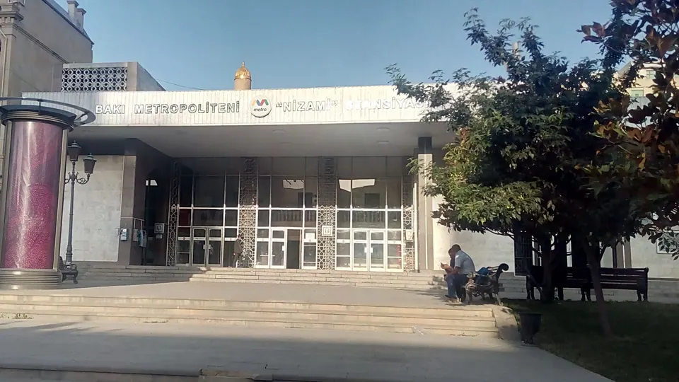 A low metro station entrance with glass doors and the station name above, with steps, a small forecourt, a tree and benches in front.