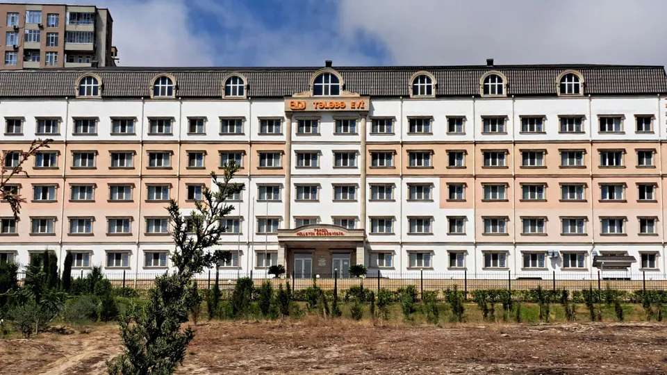 A multi-storey student residence with a white and pale peach facade and a dark roof, with a fence, young trees and open ground in front.