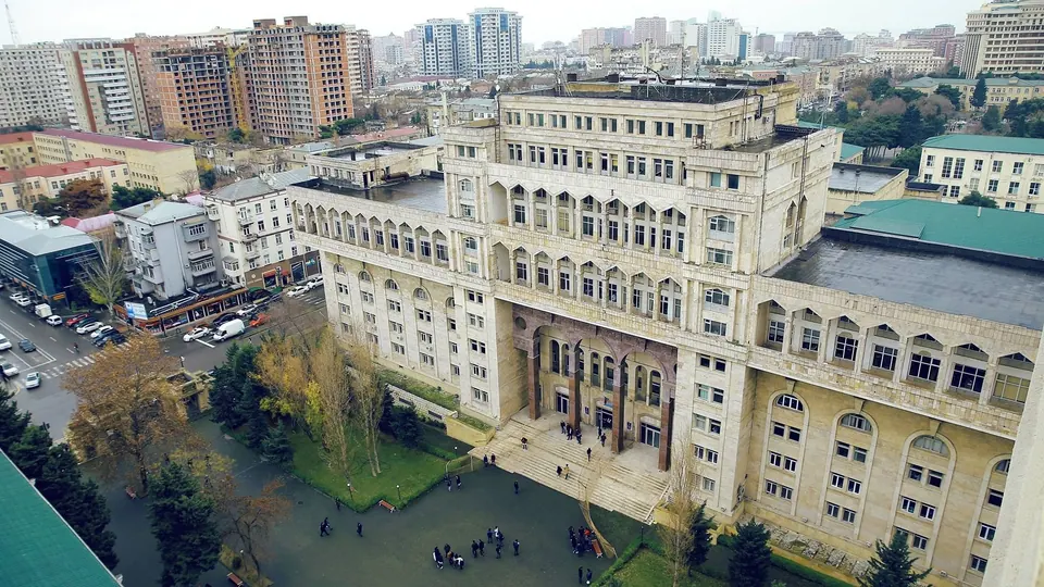 A large multi-storey stone building with arches at its entrance, a courtyard and trees in front, and residential blocks of Baku behind.